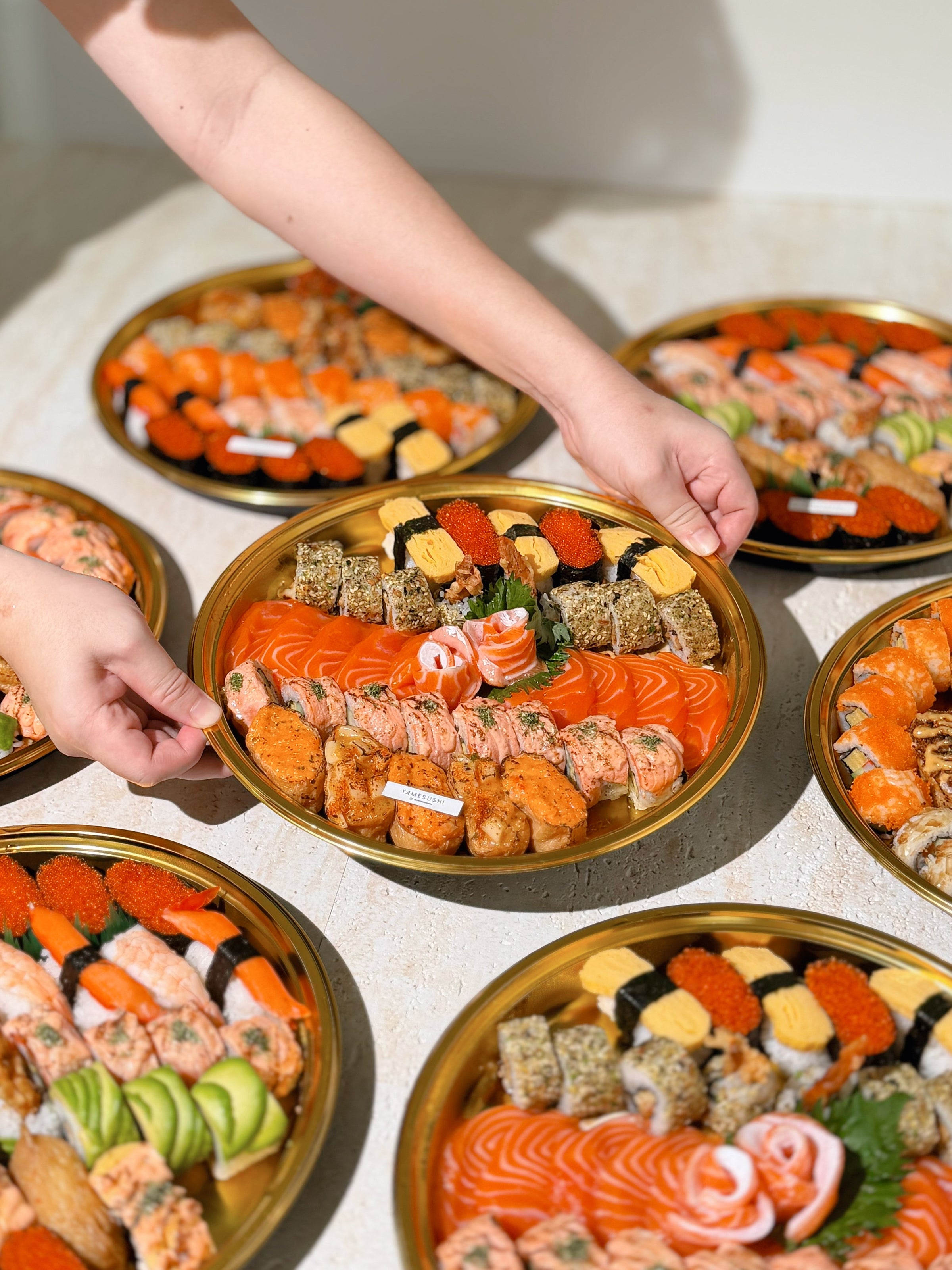 A gold-plated round platter filled with assorted sushi and sashimi, held by two hands over a light-colored table. Multiple similar platters are arranged around it.