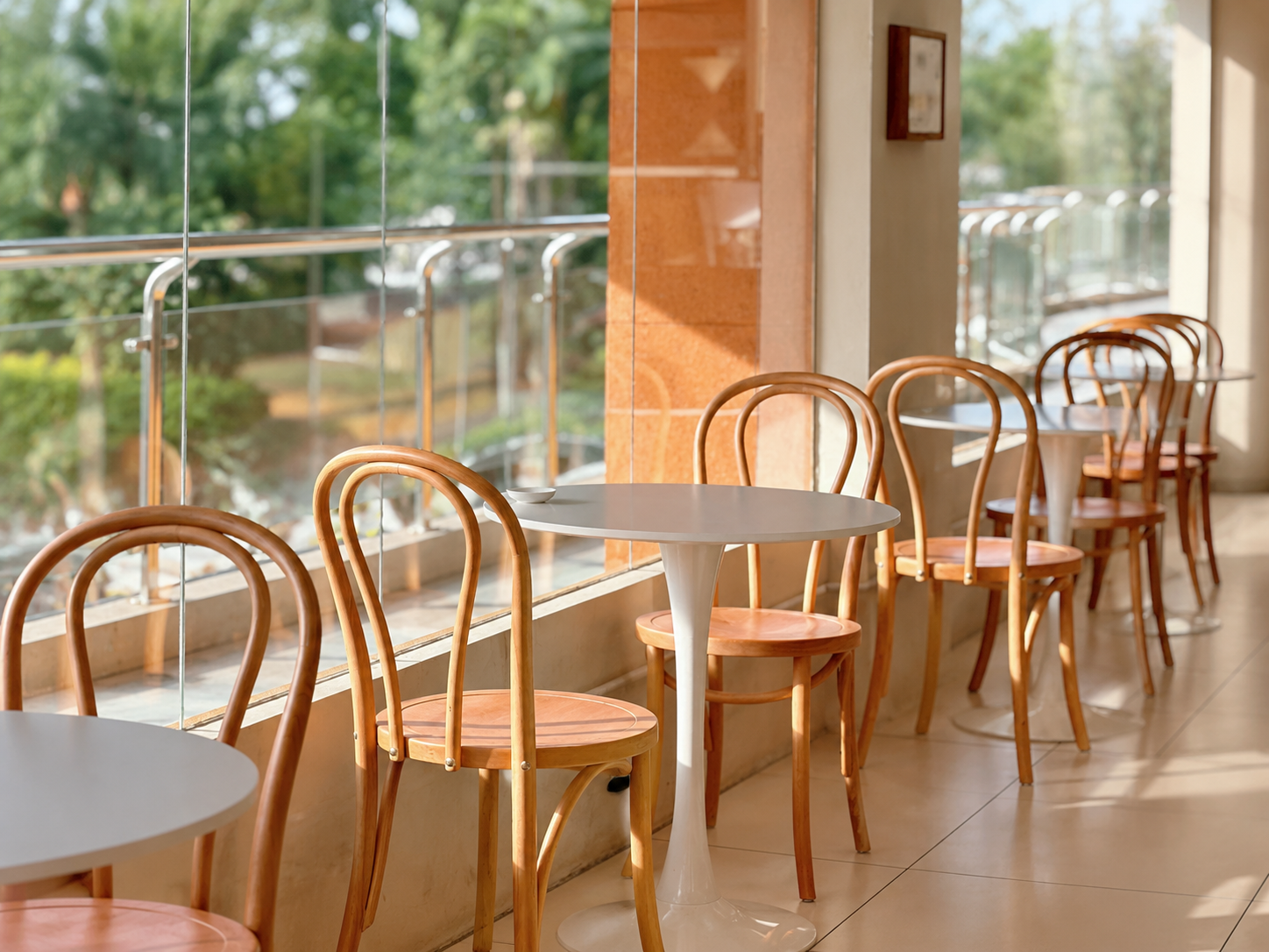 A row of round white tables and bentwood chairs with light wood seats in a bright indoor space with large windows and a glass railing.