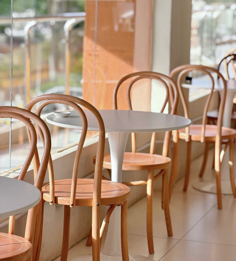 A row of light wood bentwood chairs paired with round white tables in a bright, modern cafe setting with a large window.